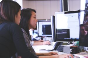 Two women with black hair look on as they read what appears on a computer monitor.