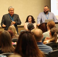 Three people stand or sit at the front of a room filled with people and talk to them at a conference.