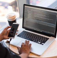 A woman sits at a desk at a laptop computer and uses the touchpad as she holds a phone in her other hand.