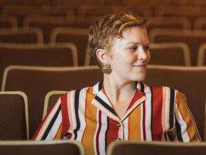 Hannah has a colorful, striped shirt on and short, cropped blonde hair. She sits in an auditorium.